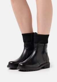 Schwarze Lederknöchelschuhe mit einem gerippten schwarzen Strickbund, seitlichem Reißverschluss und kontrastierendem roten Nähten entlang der Sohle. Strukturierte Oberflächenbeschaffenheit.