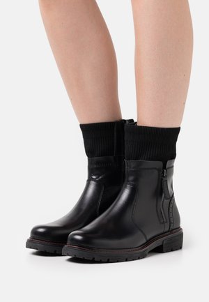 Black leather ankle boots with a ribbed black knit cuff, side zipper, and contrasting red stitching along the sole. Textured surface finish.