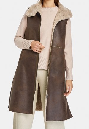 Femme portant un gilet en cuir marron foncé sans manches avec col en peau lainée sur un pull beige et un pantalon crème.