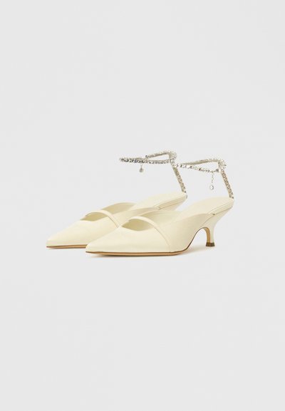 Pointed-toe cream slingback shoes made of smooth fabric, featuring a thin ankle strap with a crystal accent and a kitten heel.