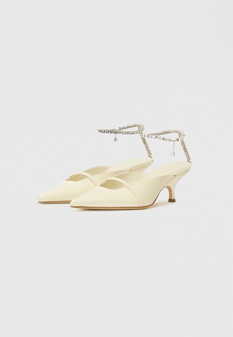 Zapatos de tacón bajo en tono crema con punta afilada, confeccionados en tela suave, que cuentan con una delgada correa para el tobillo con un acento de cristal.