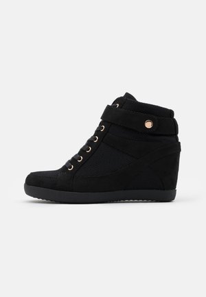 Zapatilla cuña negra con cordones en la parte frontal, correa en el tobillo con botón dorado, y textura de malla y ante, mostrada de perfil sobre fondo blanco.