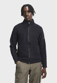 Fleecejakke i mørk navy med høj krave, fuld lynlås og sidelommer; har en struktureret overflade og subtil logoaccent.