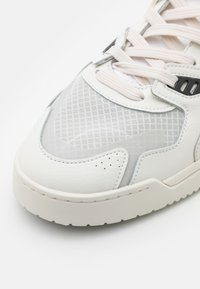 Zapatilla blanca con parte superior de cuero texturizado, paneles de malla y suela de goma. Presenta acentos grises y cordones de color claro.