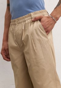 Beige, pleated trousers with an elastic waistband and side pockets. Made from smooth fabric, features a relaxed fit and clean lines.