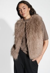 Frau mit langen dunklen Haaren, die eine flauschige braune Fellweste über einem braunen kurzärmeligen Pullover und schwarzen Hosen trägt. Sie posiert mit einer Hand in der Tasche.