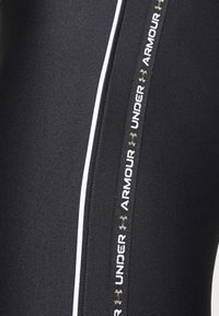 Svarta träningsleggings med en slät texture, vita sidopaneler och en framträdande silver- och vit "Under Armour"-logotyp.