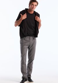 T-shirt en coton noir avec logo en relief, jean en tissu extensible gris et sneakers noirs avec semelle blanche. Le modèle pose avec une veste drapée sur les épaules.