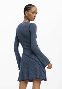 Modré šaty s dlhými rukávmi z textúrovaného materiálu, s asymetrickým lemom a kontrastným prešívaním. Disponujú priliehavým živôtikom a rozšírenou sukňou.