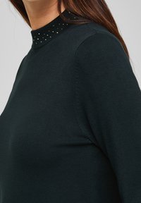 Pull noir à col montant orné de petits clous argentés. Tissu doux et texturé avec une coupe ajustée et manches longues.