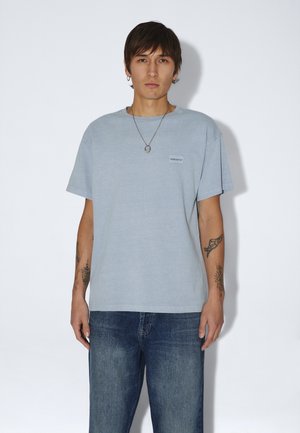 Ung person med kort mørkt hår iført lyseblå t-shirt, jeans, sølvhalskæde og viser tatoveringer på begge underarme mod en ensfarvet baggrund.