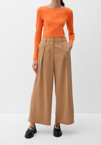 Maglietta arancione a maniche lunghe abbinata a pantaloni beige a gamba larga, con tasche laterali e mocassini neri con dettagli a catena.