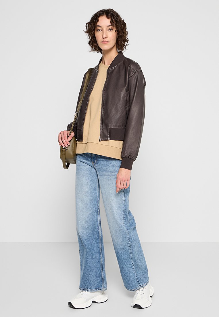 Chaqueta bomber de cuero marrón con puños acanalados, sudadera beige, jeans de pierna ancha de color azul claro y zapatillas blancas. Llevando un bolso pequeño verde.