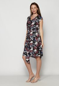 Rochie florală neagră cu modele roșii, albe și albastre, cu mâneci scurte, talie legată și tiv asimetric; asociată cu sandale cu toc transparent.