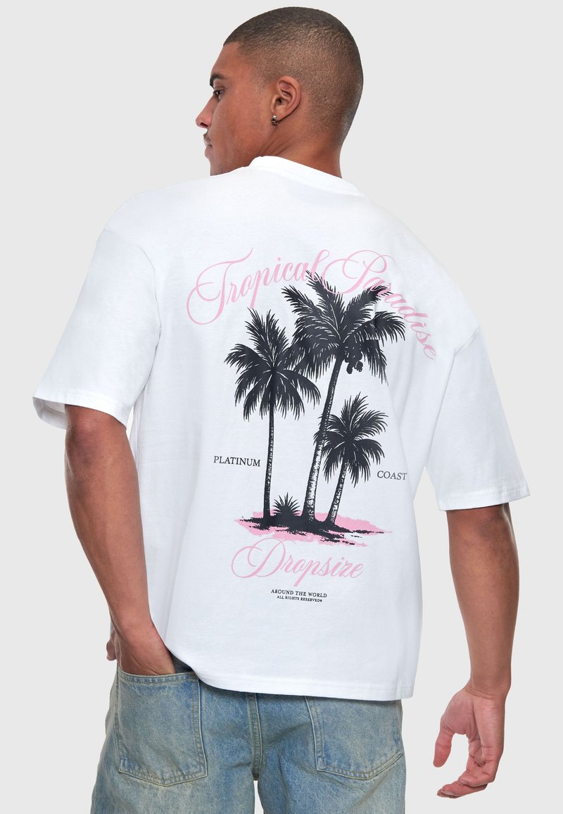 Homme portant un t-shirt blanc avec un motif de palmier noir et un texte rose indiquant « Tropical Paradise » et « Dropsize » dans le dos.