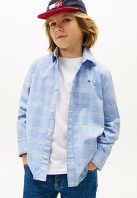Camicia azzurra a quadretti con dettagli bianchi, indossata sopra una t-shirt bianca. Abbinata a jeans di denim blu e un berretto blu navy.