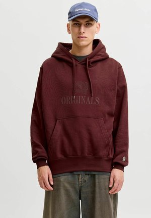 Young man wearing a maroon hoodie with front pocket, grey jeans, and a blue cap, standing against a plain light background.
