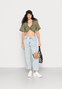 Camisa curta verde oliva com frente torcida, jeans rasgados azuis claros, ténis brancos e um chapéu preto tipo balde a segurar uma bolsa estampada.