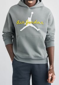 Personne atteinte de vitiligo portant un sweat à capuche gris Air Jordan avec une poche kangourou et un pantalon bleu marine, se tenant devant un fond blanc.