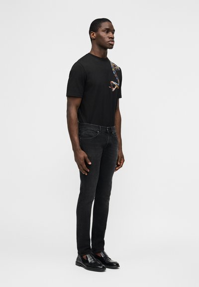 Homme debout portant un t-shirt noir avec un motif de serpent, un jean noir slim et des mocassins en cuir noir, sur un fond uni.