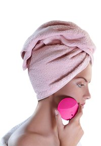 Różowe silikonowe urządzenie do oczyszczania twarzy trzymane przy twarzy, z miękkim różowym ręcznikiem owiniętym wokół głowy w stylu turbanu.