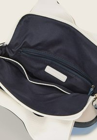 Unutrašnjost bijele teksturirane torbice s mornarsko plavim oblogom, sadrži zip džep i više otvorenih pregrada, vidljiv detalj kontrastnog rubnog obruba.