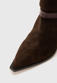 Brown suede ankle boot with a pointed toe and a dark brown leather accent strap, featuring a textured surface and a black sole.