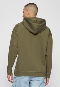 Olíva zöld kapucnis pulóver húzózsinóros kapucnival, laza szabással és látható varratokkal. A háta egyszínű textúrát mutat, grafikák nélkül.