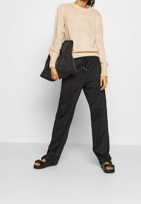 Beige neulepusero, mustat elastiset vyötäröhousut, musta laukku pidetty yhdessä kädessä ja mustat sandaalit solkikoristein. Näkyvissä sileät tekstuurit.