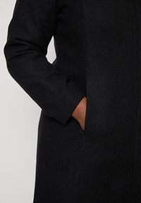 Person mit mittelbrauner Haut, der einen schwarzen Langarmmantel trägt und deren rechte Hand in die Seitentasche gesteckt ist.