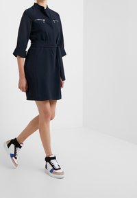 Granatowa sukienka z paskiem w talii, zapinanymi kieszeniami na piersiach i rękawami do łokcia, zestawiona z kolorowymi sneakersami typu high-top.