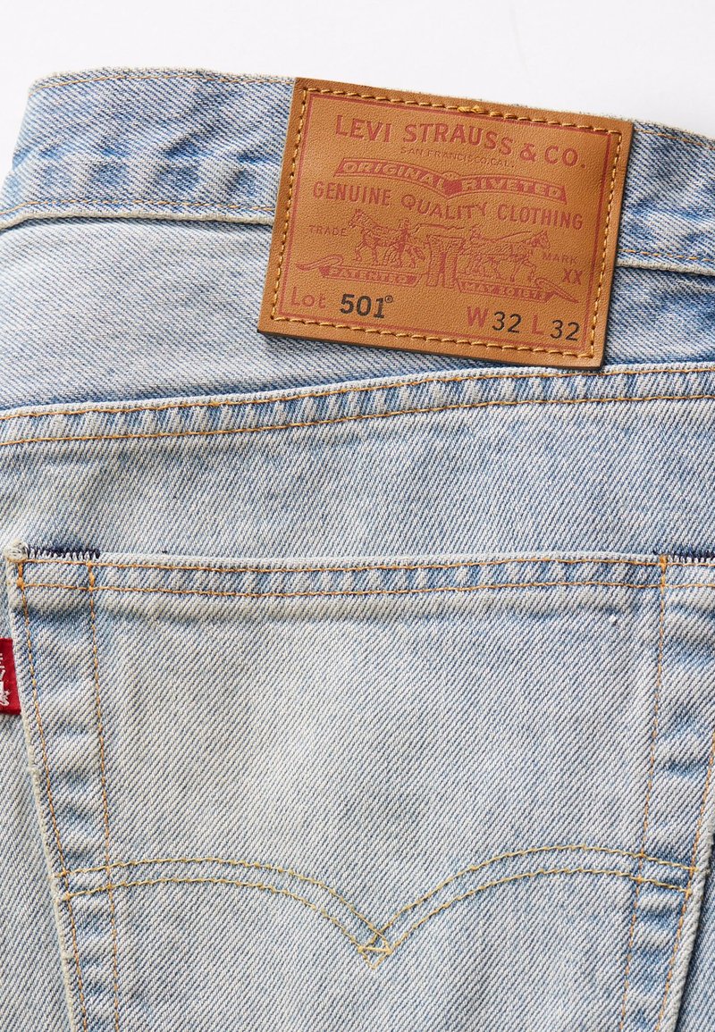Jeans in denim azzurro chiaro con etichetta in pelle marrone recante il nome del marchio in rilievo, il numero del lotto e le informazioni sulle taglie. Cuciture a contrasto visibili.