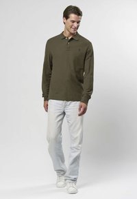 Långärmad olivgrön pikétröja med krage och svart logotyp, kombinerad med ljusblå jeans och vita sneakers på en modell som står mot en ljus bakgrund.