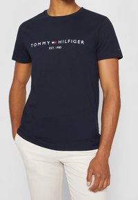Camiseta de algodón azul marino con cuello redondo, que presenta el texto y el logo de “TOMMY HILFIGER” impresos en blanco en el pecho.