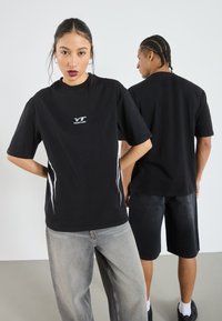 Svart oversize T-shirt i bomull, med en vit logotyp på bröstet och vita sidostycken. Modellen har den matchat med ljusgrå jeans.
