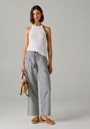 Naine, kes kannab valget varrukateta kootud topi, triibulisi laia säärega pükse ja pruunid sandaale, hoides käes väikest nahkrihmadega kootud kotti.