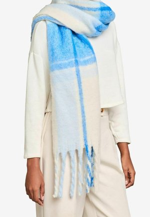 Écharpe douce à carreaux bleue et blanche avec franges, enroulée autour du cou, portée sur un haut crème à manches longues et un pantalon beige.