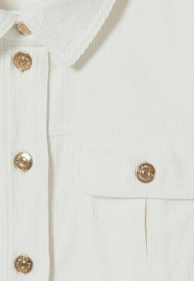 Camicia a coste bianca con colletto classico, caratterizzata da bottoni dorati e dettaglio sulla tasca. La texture presenta evidenti righe verticali.