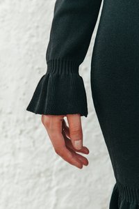 Schwarzer gestrickter Pulloverärmeln mit strukturiertem Rippmuster und weit ausgestelltem Bündchen. Sichtbare Hand, die gegen einen strukturierten weißen Hintergrund ruht.