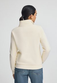 Felpa color crema con colletto alto, texture morbida e orlo a coste, abbinata a jeans in denim blu. Nessun motivo visibile o accessorio.