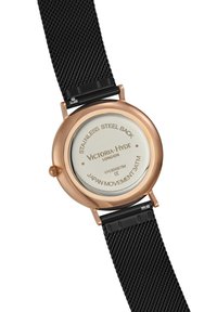 Okrągły zegarek z polerowaną kopertą w kolorze różowego złota, szklaną tylną częścią, czarnym meshowym paskiem oraz grawerowanymi detalami, w tym "Ruch japoński 3 ATM" i nazwą marki.