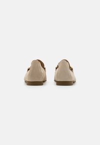Beige suède loafers met een lage bruine zool, voorzien van een open achterkant en gouden hardware-accenten aan de zijkanten, een gladde textuur en een ronde vorm.