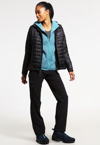 Schwarze gesteppte Jacke über einem türkisfarbenen Fleece-Reißverschluss, kombiniert mit schwarzen Hosen und blauen Wanderschuhen, die ein Reißverschlussdesign und eine gemütliche Textur aufweisen.