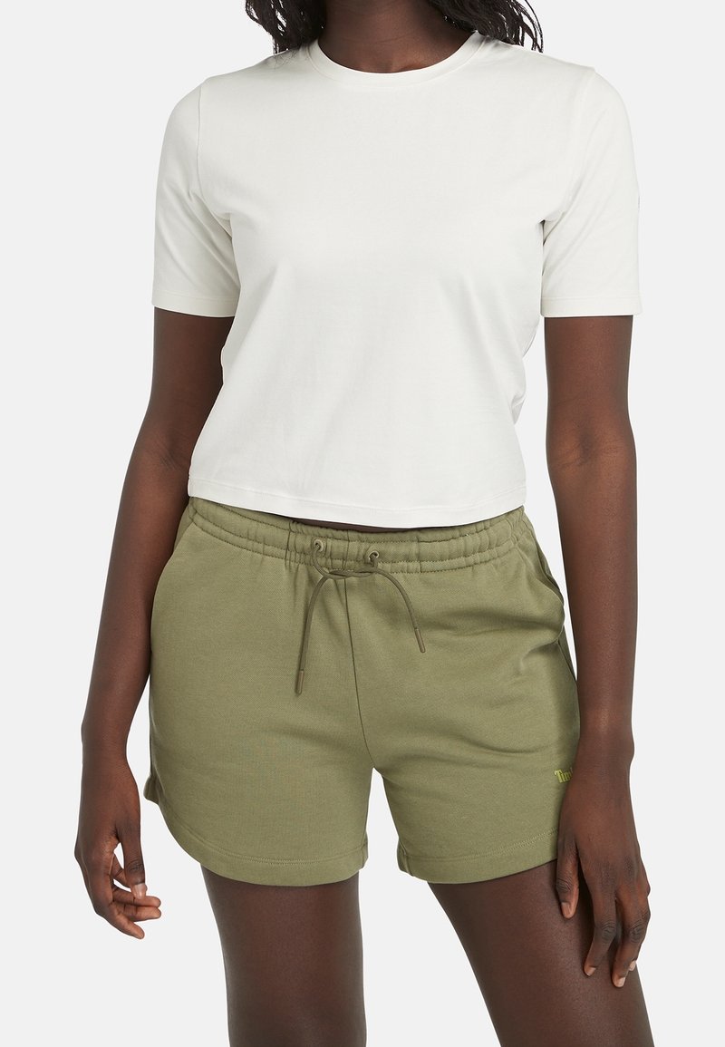 Femme portant un haut blanc uni à manches courtes et un short vert olive avec cordon, debout devant un fond neutre.