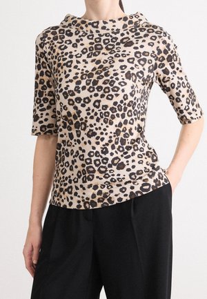 Kvinde iført en beige leopardprint kortærmet top kombineret med sorte højtaljede bukser med den ene hånd i lommen.