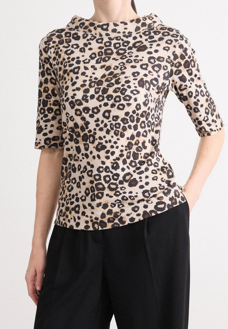 Femme portant un haut beige à manches courtes imprimé léopard associé à un pantalon noir taille haute, une main dans la poche.
