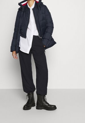 Persoon die een marineblauwe donsjas draagt, een wit overhemd met knopen, zwarte broek en zwarte vetercombatlaarzen, staand tegen een effen achtergrond.