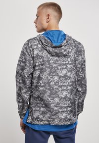 Jachetă de camuflaj în nuanțe de gri, cu glugă și căptușeală albastră. Materialul are o textură netedă și manșete elastice.