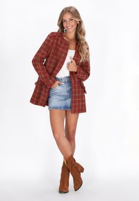 Cappotto in tweed a scacchi rossi e verdi con una vestibilità dritta, abbinato a una maglietta bianca e una gonna in denim. Stivali in suede marroni completano l'outfit.