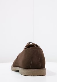 Chaussure en daim marron à faible talon, forme arrondie et détails cousus. L'arrière présente une texture douce et une semelle grise subtile.